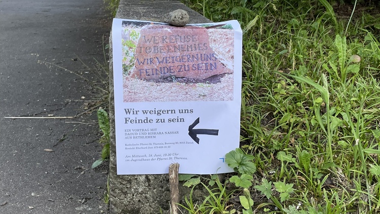 Am Wegrand weist ein Plakat auf die Veranstaltung in der Pfarrei St. Theresia in Zürich hin.
