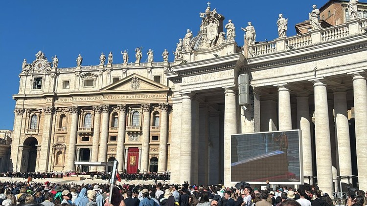Jugendliche am Petersplatz vor einem Screen, auf dem der Sarg von Papst Franziskus zu sehen ist.