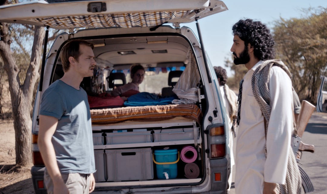 Filmstill: David unterhält sich vor dem Reisebus mit einem bewaffneten Mann, Daniela sitzt am Steuer