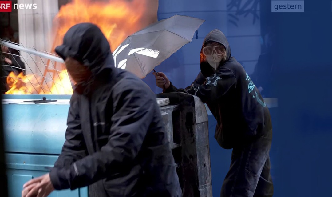 Das Foto zeigt zwei vermummte Demonstrierende vor einem brennenden Container. Einer der Vermummten trägt fünfzackige Sterne auf dem Ärmel.