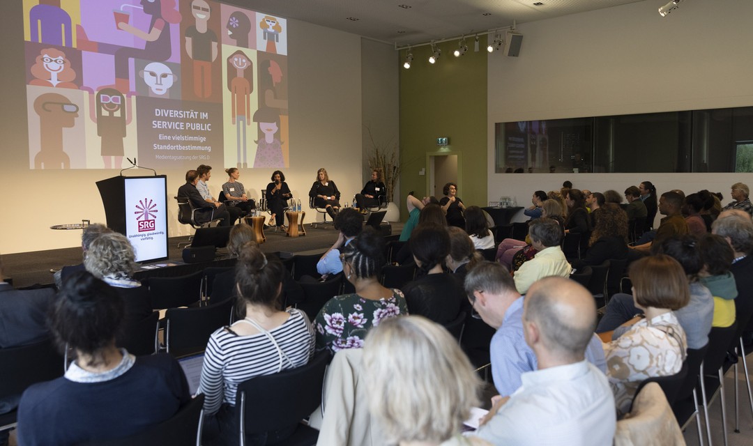Das Publikum schaut auf die Bühne, wo eine Diskussion mit fünf Expert:innen stattfindet