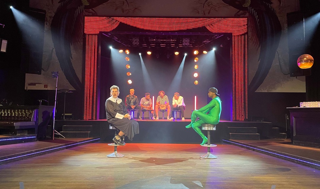 Auf einer Bühne eines Nachtclubs sitzten mehrere Menschen.