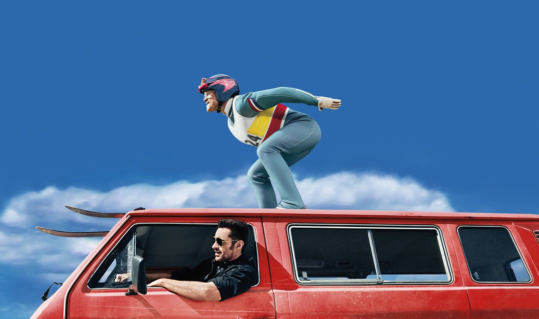 Taron Eggerton in Skispringmontur auf Dach von Auto, das von Hugh Jackman gefahren wird. Ausschnitt «Eddie the Eagle»