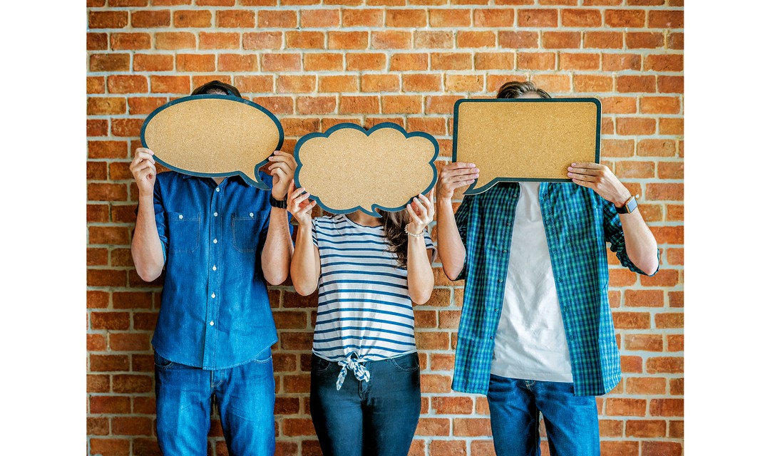 Drei Menschen stehen vor einer Backsteinwand und halten sich Sprechblasen aus Karton vors Gesicht