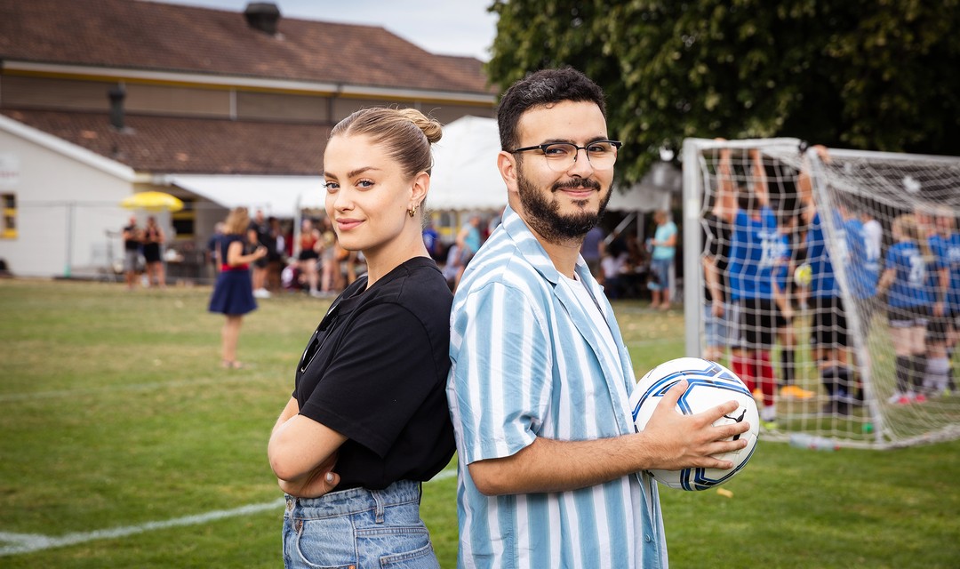 Jennifer Bosshard (SRF) und Yacine Nemra (RTS) stehen zusammen vor einem Fussballplatz. Yacine Nemra hält einen Fussball in den Händen.