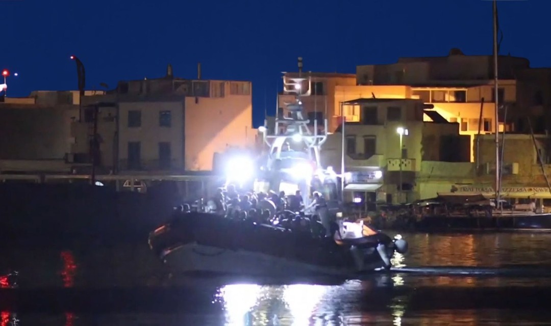 Das Foto zeigt ein Boot voller Flüchtlinge, das in einen kleinen Hafen bei Nacht einfährt. Das Boot ist mit hellen Scheinwerfern beleuchtet.