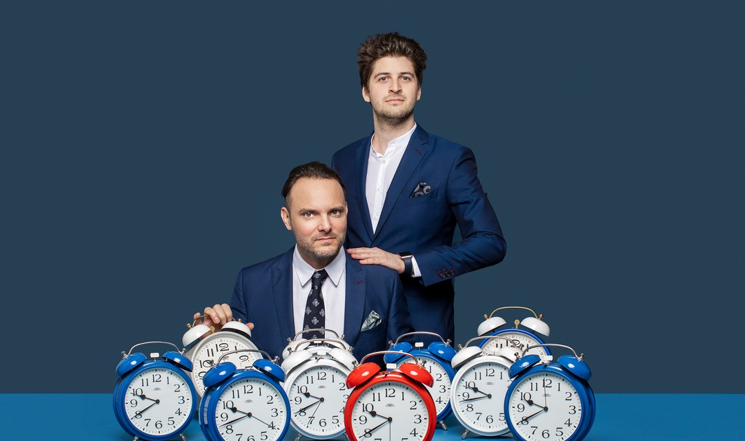Dominic Deville und Patrick Karpiczenko stehen hinter einem Tisch voller analoger Wecker.
