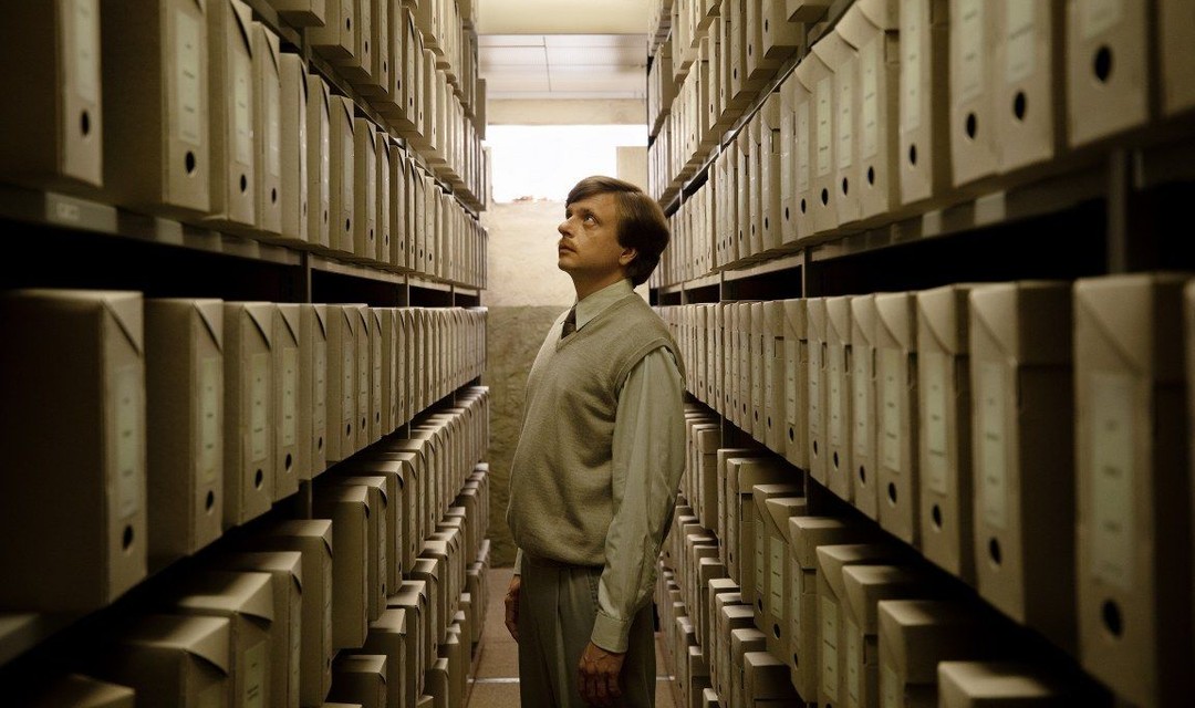 Der Polizeibeamte Viktor steht in einem riesigen Archiv, neben ihm hat es Regale mit Ordnern