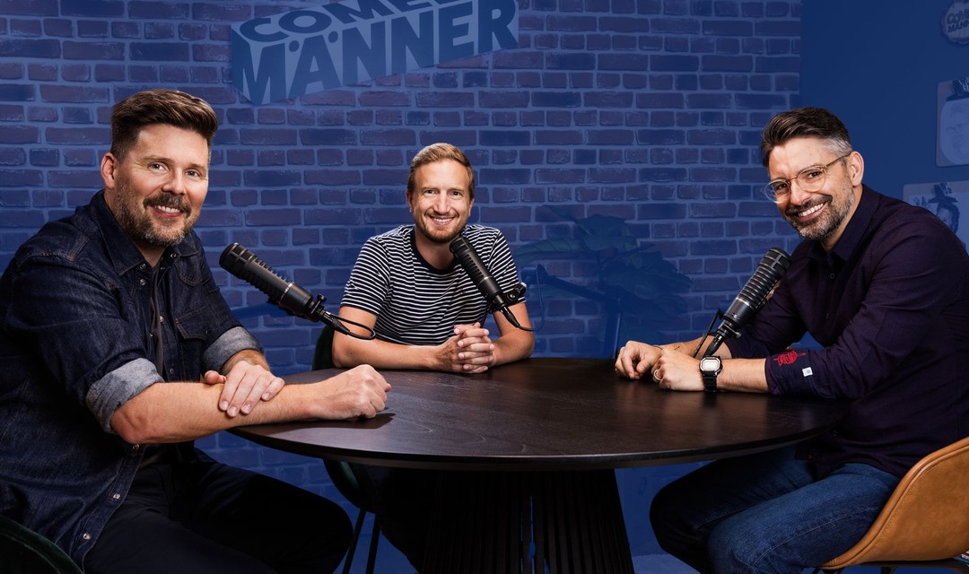 Das Foto zeigt die drei Comedymänner. Sie sitzen an einem runden Tisch und haben je ein Mikrofon vor sich.